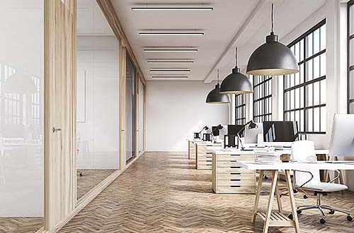 Front view of an office interior with a row of dark wood tables standing under large windows. Massive ceiling lamps. Computers.